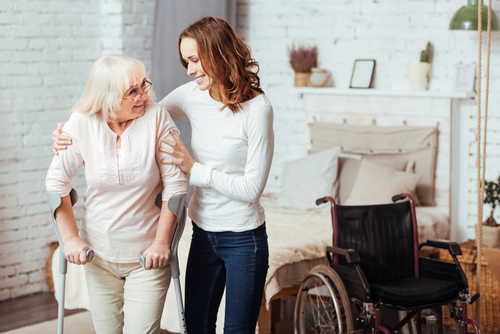 Daughter assisting her elderly mother with walking crutches standing next to wheelchair