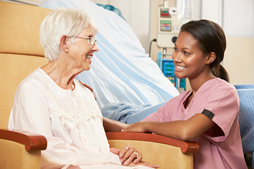 Young woman in light pink scrubs knelt down smiling at elderly woman wearing a night gown sitting in chair